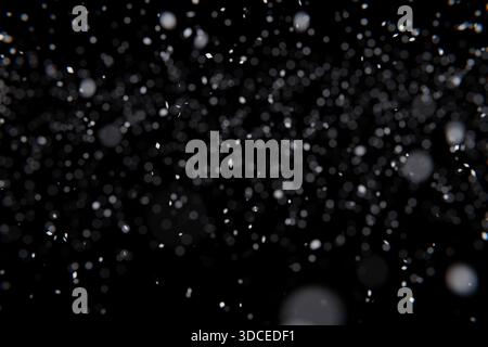 Abstrakter Schnee, der auf schwarzen Hintergrund fällt. Verschwommenes weißes Bokeh und scharfe Partikel erzeugen eine dynamische, dunkle Winterüberlagerung. Ideal für Urlaubsdesigns, Stockfoto