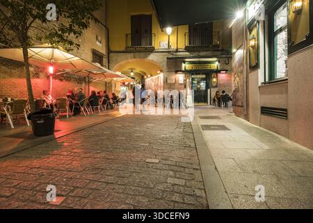 Madrid, Spanien - 09. November 2016: Blick auf ein belebtes Café, das auf Kopfsteinpflasterstraßen übergeht, die in das warme Licht der Straßenlaternen und das einladende Re getaucht sind Stockfoto