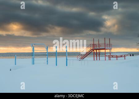 Blick auf rote Metalltreppen und blaue Metallstangen an einem schneebedeckten Strand unter einem bewölkten, goldgefärbten Himmel, Ostsee, Parnu, Estland. Stockfoto