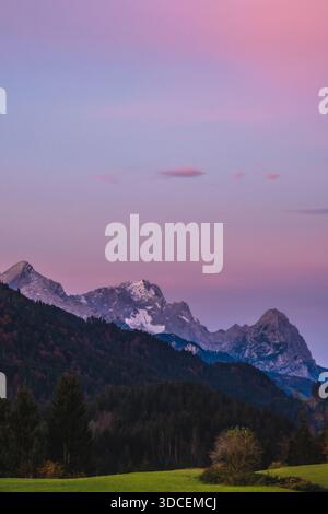 Der Blick auf eine ruhige Landschaft entfaltet sich mit grünen Feldern, die auf einen dunklen, bewaldeten Hügel und schneebedeckte Berge unter einem pastellfarbenen Himmel treffen, Krun, Deutschland. Stockfoto