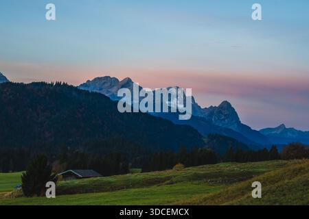 Blick auf eine ruhige Landschaft mit sanften grünen Hügeln, die auf einen dunklen Wald und majestätische Berge unter einem pastellfarbenen Himmel in der Abenddämmerung treffen, Krun, Deutschland. Stockfoto