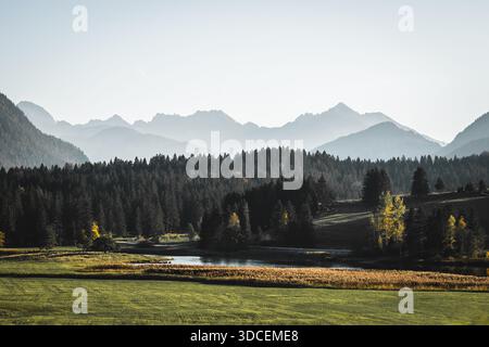Blick auf einen ruhigen See, der den umliegenden dunklen Wald und die fernen, trüben Berge unter hellem Himmel reflektiert, Krun, Bayern, Deutschland. Stockfoto