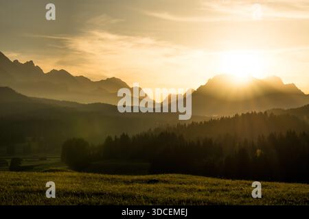Der Blick auf goldenes Licht taucht über die zerklüftete Bergkette und wirft lange Schatten über den üppigen Wald und die Wiese in Krun, Bayern, Deutschland. Stockfoto