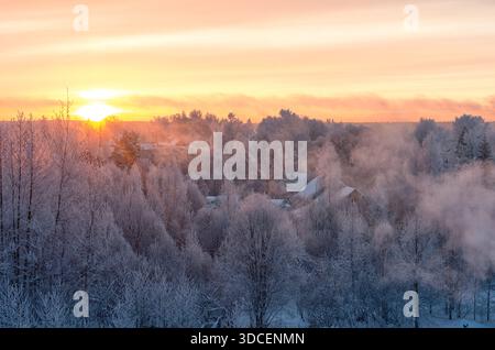 Malerisches Winterdorf bei Sonnenaufgang mit frostigen Bäumen und Dampf, der aus Häusern in rosa und orange Himmel steigt, Luftlandschaft, die kalte Morgenstimmung einfängt Stockfoto