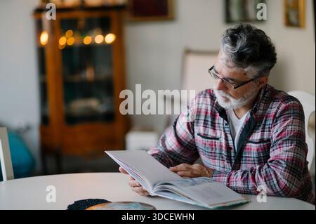 Senior Mann liest ein Buch zu Hause in einem gemütlichen Wohnzimmer Stockfoto