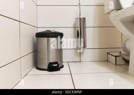Mülleimer aus Edelstahl mit Fußpedal auf dem Boden und einer wandmontierten Toilettenbürste in einem Badezimmer mit großen hellen Keramikfliesen Stockfoto