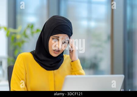 Muslimische Geschäftsfrau, die einen Hijab trägt und an einem Schreibtisch sitzt, unter schweren Kopfschmerzen leidet, ihren Tempel hält, während sie auf einen Laptop-Bildschirm blickt, betont arbeitsbedingten Stress und Müdigkeit Stockfoto