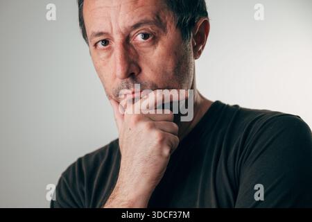 Porträt eines Mannes mittleren Alters mit kurzen dunklen Haaren und Stoppeln, der nachdenklich mit Hand am Kinn vor neutralgrauem Hintergrund blickt. Reflexion, wor Stockfoto