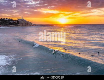 Aus der Vogelperspektive auf Surfer, die die Wellen bei Sonnenuntergang in der Nähe der historischen Altstadt von Jaffa mit ihren alten Gebäuden vor dem farbenfrohen Himmel fangen, Stockfoto