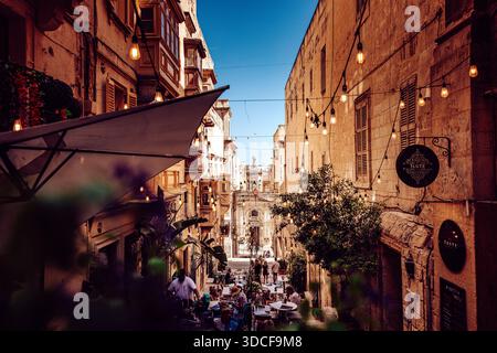 [Valletta], [Malta] - 03. August 2024: Blick auf eine enge Straßenlandschaft in Valletta, Malta, wo warme Steinbauten eine ferne Kirche säumen, beleuchtet Stockfoto