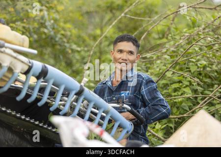 Phu Tho, Vietnam - 15. Oktober 2025: Der Blick auf einen Mann in einem blau karierten Hemd lächelt inmitten des dichten grünen Laubs, ein starker Kontrast zum hellblauen mac Stockfoto