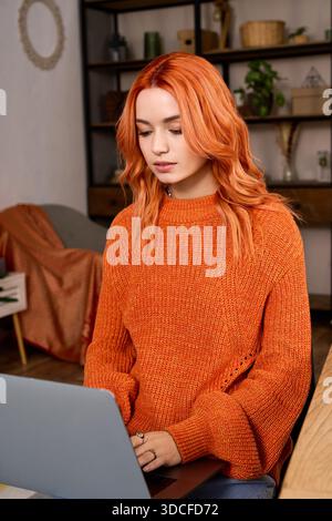 Die junge Frau mit rotem Haar genießt einen gemütlichen Wintertag zu Hause und arbeitet an ihrem Laptop. Stockfoto