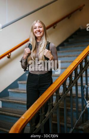 Lächelndes Porträt einer blonden Studentin, die einen Rucksack trägt, der auf einer Treppe im Universitätsgebäude steht Stockfoto