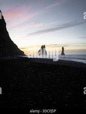 Blick auf den schroffen schwarzen Sand, der auf die schaumige Brandung unter dramatischen Klippen trifft, und die riesigen Meeresstapel, die von den sanften Tönen der Dämmerung gemalt sind, Reynisfjara, Island Stockfoto