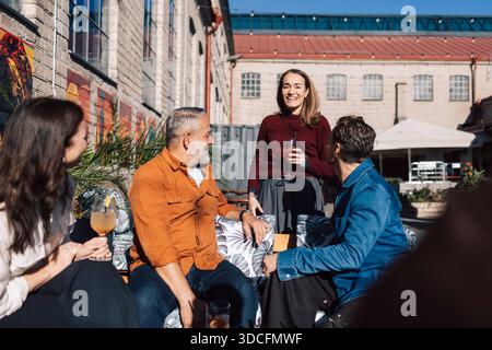 Lächelnde Frau mit Freunden, die Getränke genießen, während sie an sonnigen Tagen draußen an der Bar sitzt Stockfoto