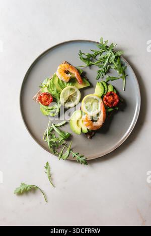 Vegetarisches Sandwich mit in Scheiben geschnittenen Avocado, sonnengetrocknete Tomaten, Garnelen, Rucola, serviert auf keramische Platte über weißem Marmor Hintergrund. Flach, Raum Stockfoto