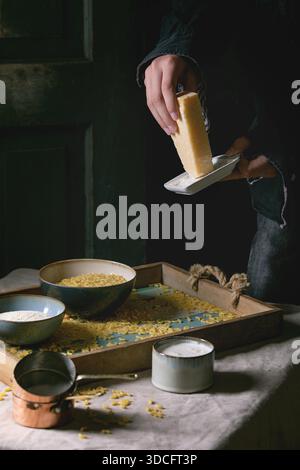 Junger Mann in schwarzem Käse. Rohe, ungekochte italienische Pasta Coquillettes zum Kochen des Abendessens mit Parmesan, Sahne und Grießmehl auf Holztablett auf Küchenwäsche Tischtuch. Dunkler, rustikaler Stil. Stockfoto
