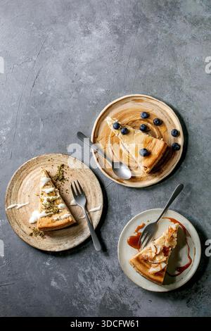 Käsekuchen mit verschiedenen Belägen und Dekor auf Keramikplatten mit Gabeln über grauem Texturhintergrund. Flaches Layout, Platz Stockfoto