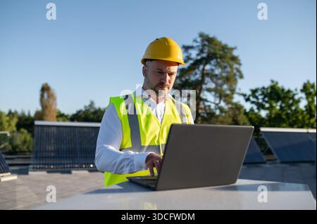 Ingenieur im Helm, der an Laptop in der Nähe von Solarpaneelen im Freien arbeitet Stockfoto