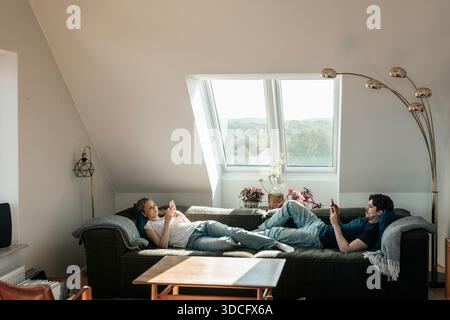 Junge Mann und Frau, die ihr Smartphone benutzen, während sie auf dem Sofa in der Wohnung liegen Stockfoto