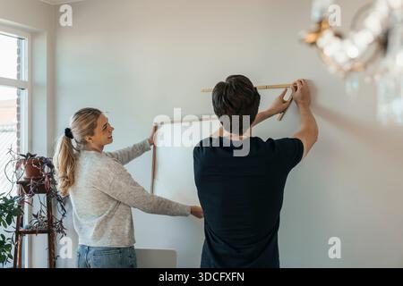 Junger Mann, der an der Wand gemessen wird, um Bilderrahmen zu hängen, während der Umsiedlung mit Freundin Stockfoto