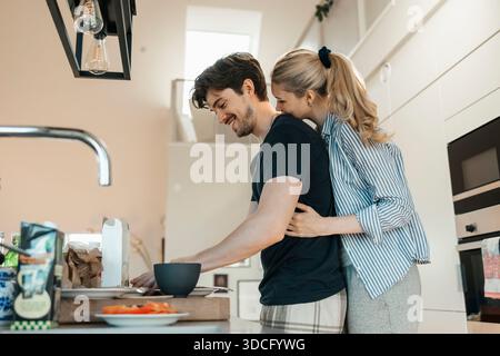 Niedriger Blickwinkel einer jungen Frau, die einen Freund umschließt, der zu Hause in der Küche frühstückt Stockfoto