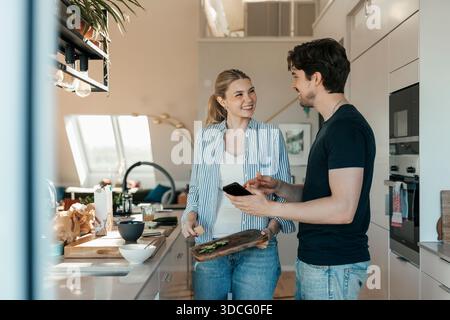 Junger Mann, der der Freundin ein Smartphone zeigt, das die Küche zu Hause reinigt Stockfoto