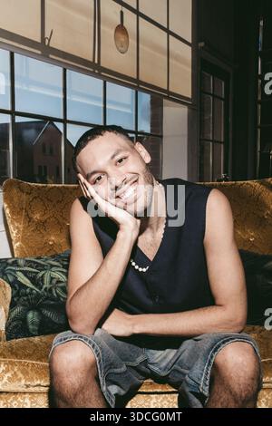 Porträt eines lächelnden jungen Mannes, der sich auf der Hand beugt, während er auf dem Sofa sitzt Stockfoto