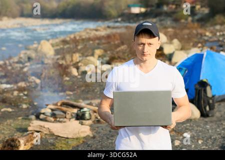 Ein ernsthaft aussehender Wohnmobil in weißem Hemd hält einen Laptop fest, hinter dem Zelt und Rucksack sichtbar sind, mit Holzfeuer und Topf auf Steinen am Herbstufer. Stockfoto