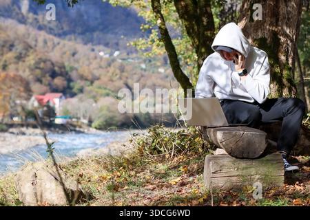 Mann in Kapuzenpullover spricht auf dem Handy, während er auf Baumstümpfen in der Nähe des Herbstwaldes sitzt, mit offenem Laptop und River vista Stockfoto