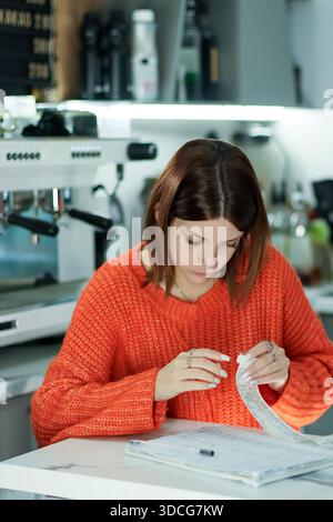Die junge Frau an der Marmortheke im Café untersucht Papierquittung mit Stift, Espresso-Maschine in unscharfem Hintergrund. Stockfoto