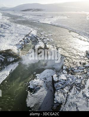 Blick aus der Vogelperspektive auf den majestätischen Wasserfall, der in den eisigen Fluss stürzt, im Kontrast zur schneebedeckten Landschaft, Vik, Western Region, IC Stockfoto