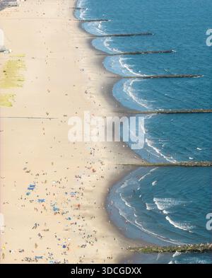 Blick aus der Vogelperspektive auf den Strand von Coney Island und die Wellenbrecher treffen sich in einem rhythmischen Tanz aus Sand und Meer, mit Menschenmassen am Ufer unter der Spätsommersonne. Stockfoto