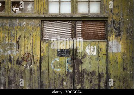Vorderansicht der alten hölzernen Industrietüren mit abblätternder Farbe, rostigen Metallgitterfenstern und sichtbaren Anzeichen von Verfall und städtischer Verlassenheit. Stockfoto