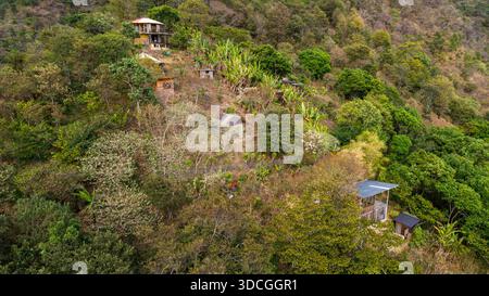 Blick aus der Vogelperspektive auf Holzkonstruktionen, eingebettet zwischen üppigen grünen Bäumen und brauner Vegetation, die den Hügel, den Atitlan-See, das Departement Sololá, Guate bedecken Stockfoto