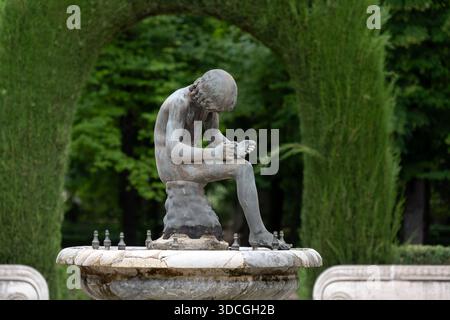Eine Bronzestatue eines sitzenden Jungen mit dem Kopf nach unten auf einem Steinbrunnen, umgeben von Grün Stockfoto