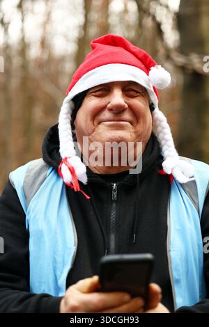 Ein lächelnder Mann im weihnachtsmann hält draußen ein Smartphone Stockfoto