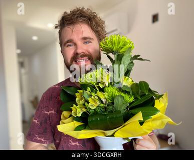 Ein lächelnder Mann, der drinnen einen Blumenstrauß hält. Geschenkte Momente, Wärme und Zuneigung, fröhlicher Ausdruck und emotionale Verbindung in hell Stockfoto