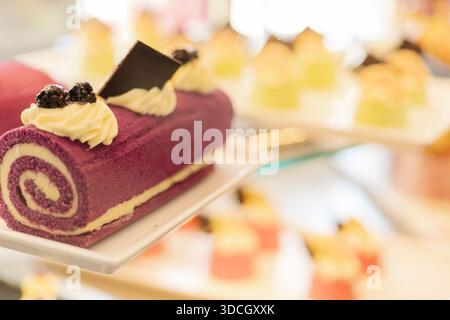 Eine Gruppe von Desserts ist auf weißen Tellern angeordnet. Ein violetter Brötchen mit Sahne und Schokolade steht im Vordergrund zwischen verschiedenen kleinen Gebäckstücken. Personen en Stockfoto