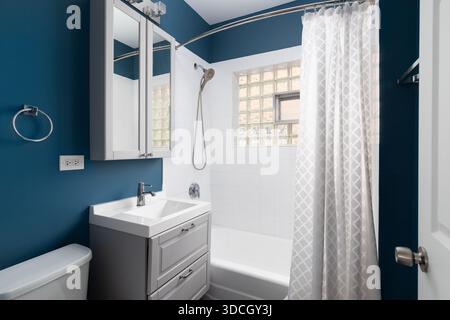 Ein blaues Badezimmer mit grauem Kosmetikbereich, weißem Waschbecken und weißen quadratischen Fliesenwänden mit Glasblockfenster. Stockfoto