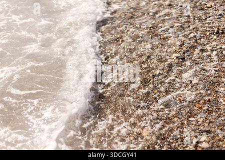 Wellen krachen auf einen Strand, der mit kleinen Steinen und Kieselsteinen bedeckt ist. Das Wasser bewegt sich ein- und auswärts und erzeugt Muster am Ufer. Sonnenlicht reflektiert das Nässe Stockfoto