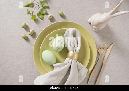 Minimaler Ostertisch mit grünen Keramikplatten, pastellfarbenen Eiern, Leinenserviette und goldenem Besteck auf neutralem Textilhintergrund. Draufsicht. Frühlingsferienkonzept. Stockfoto