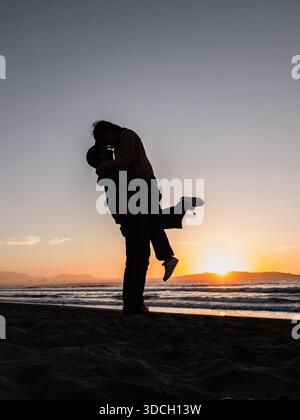 Ein Paar küsst sich am Strand bei Sonnenuntergang. Der Mann trägt eine blaue Jacke und die Frau einen gelben Pullover. Die Sonne geht hinten unter Stockfoto