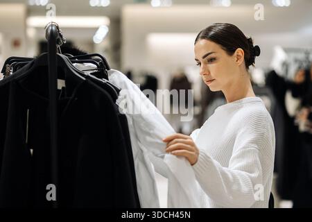 Weibliche Käuferinnen, die in einem weißen Pullover gekleidet sind, untersuchen Kleidungsstücke auf einem Regal in einem modernen Einzelhandelsumfeld und heben die Auswahl von Mode und Persönlichkeit hervor Stockfoto