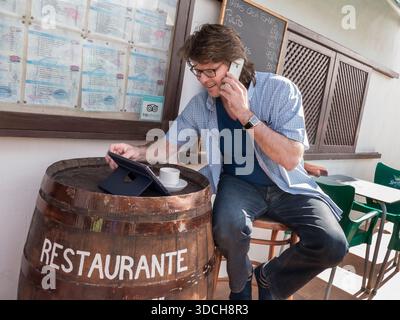 GESCHÄFTLICHE KOMMUNIKATION IM AUSLAND Reifer Mann mit seinem iPhone Smartphone und iPad Air Smart Tablet Computer im Ausland in der spanischen Restaurant Bar im Freien, mit Fassplatte als rustikaler Tisch. Lanzarote Spanien Stockfoto