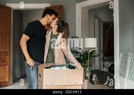 Frau, die Mann küsst, während sie in der Nähe von Pappkarton in einem neuen Zuhause steht Stockfoto