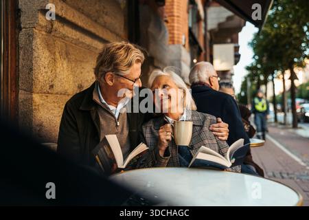 Lächelnder älterer Mann, der ein Buch hält und eine Frau umschließt, während er im Straßencafé sitzt Stockfoto