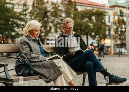 Ein älteres Paar liest ein Buch, während er auf der Bank in der Stadtstraße sitzt Stockfoto