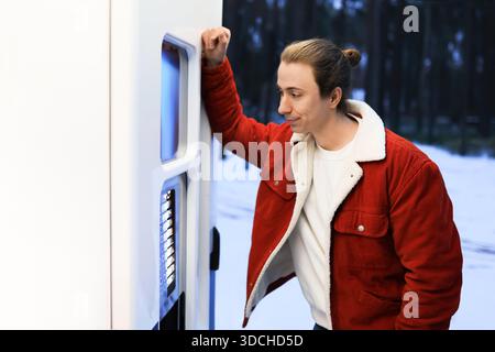 Junger Mann in roter Jacke, der sich an einem Automaten in einem verschneiten Winterpark lehnt und auf ein heißes Getränk wartet. Hochwertige Fotos Stockfoto
