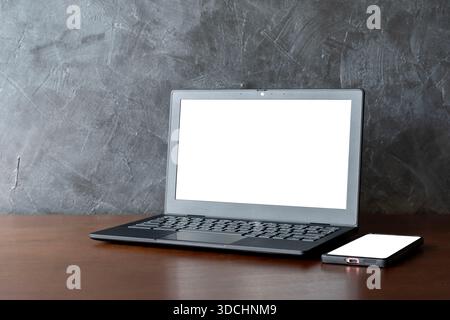 Moderner Arbeitsbereich mit offenem Laptop und Smartphone, auf dem leere weiße Bildschirme auf dem Holztisch vor einer grauen strukturierten Wand stehen. Perfekt für Mockups, Anzeigen, Web Stockfoto